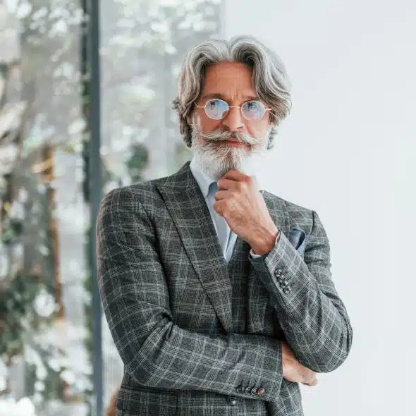 A distinguished-looking man with grey hair and beard, wearing a plaid suit and round glasses, stands with his hand on his chin.