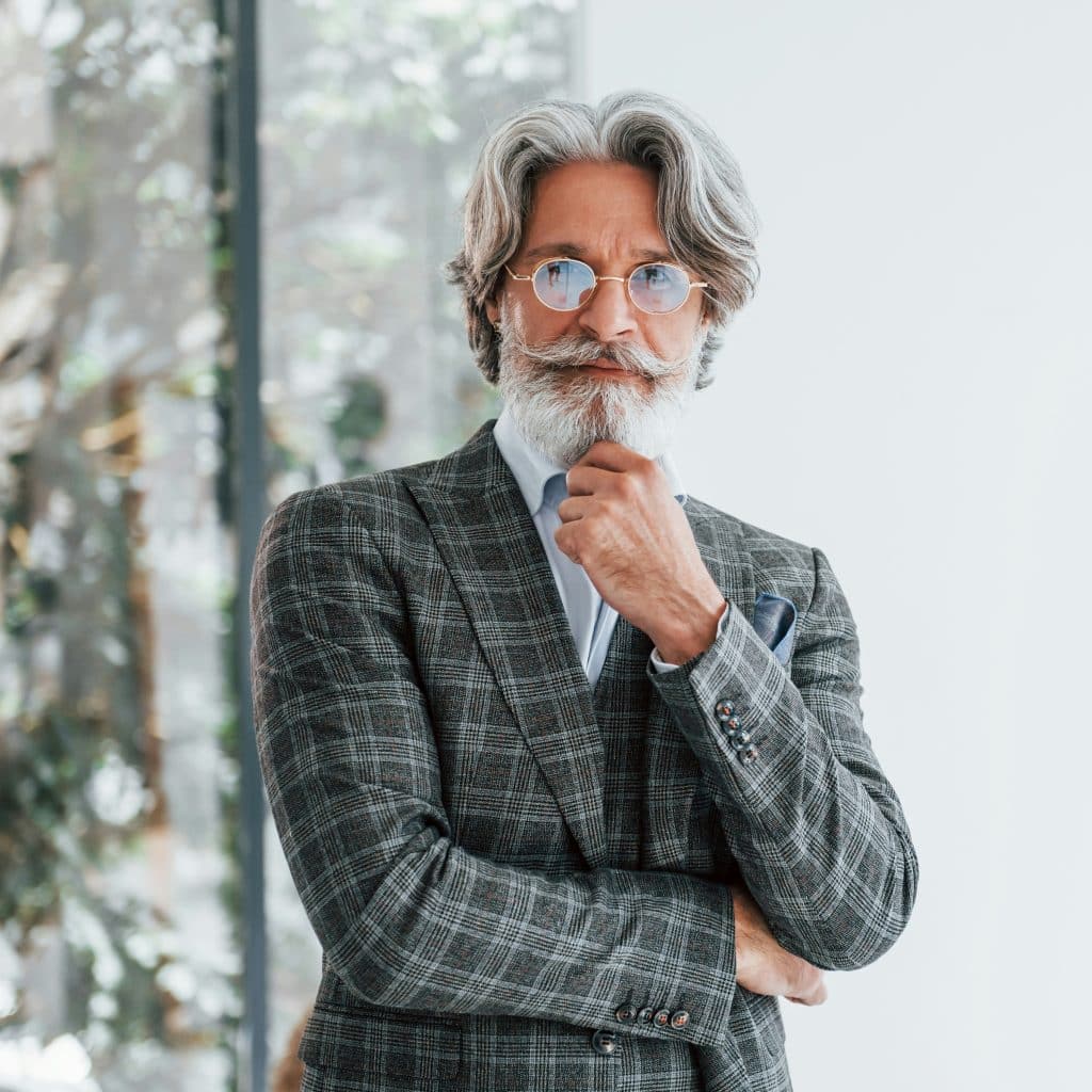 A distinguished-looking man with grey hair and beard, wearing a plaid suit and round glasses, stands with his hand on his chin.