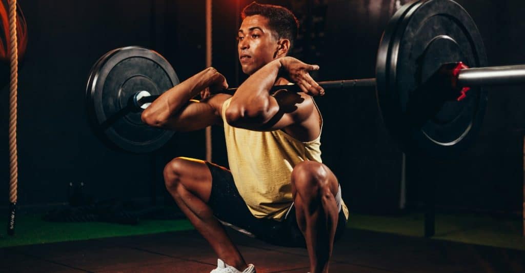 A man in a yellow tank top and black shorts is performing a deep squat with a heavy barbell resting across his upper back in a gym.