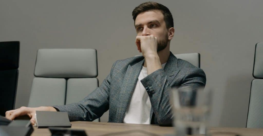 A man in a grey blazer and white t-shirt sitting at a conference table, with his hand on his chin, looking contemplative.