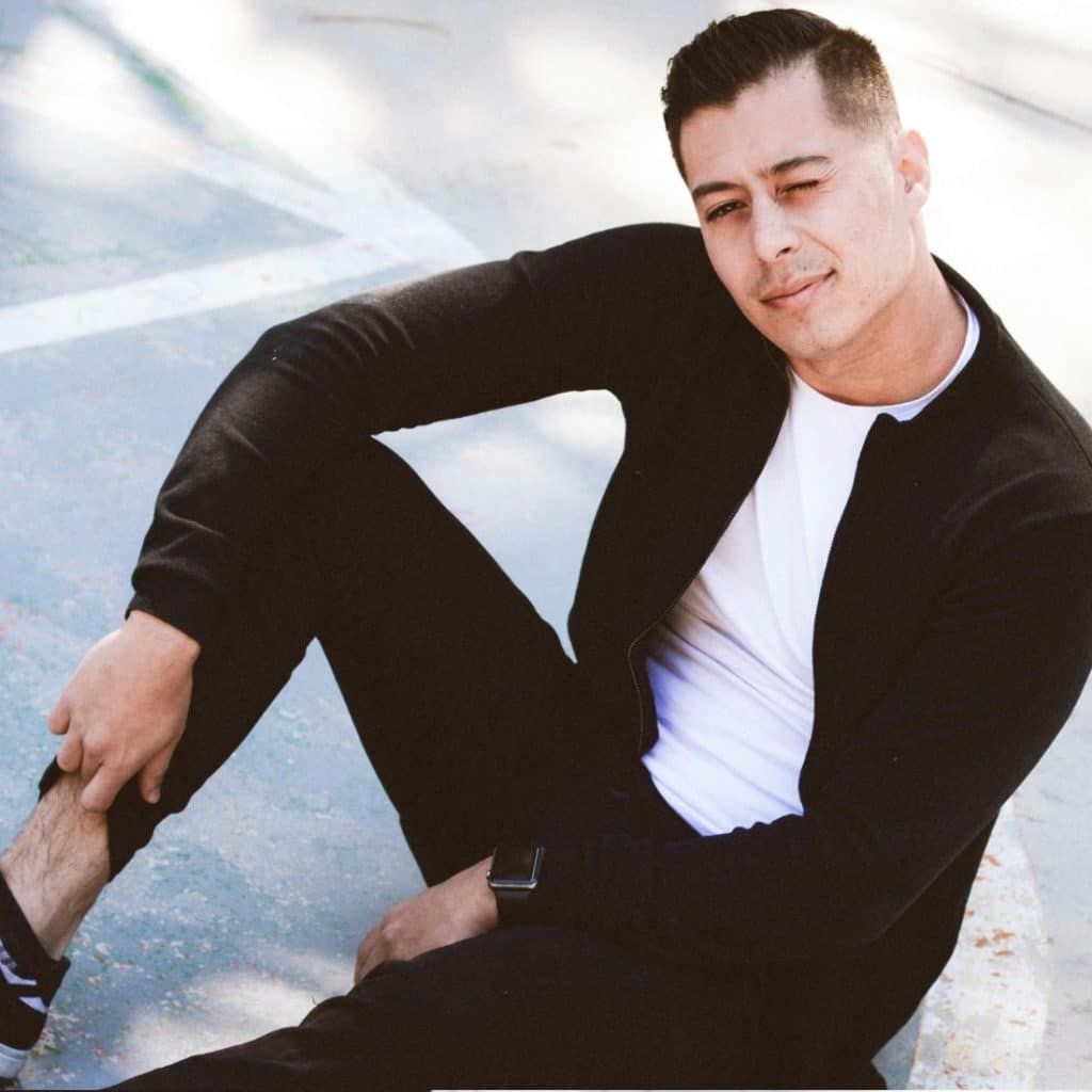 A man sitting on a basketball court, wearing a black jacket, white t-shirt, black pants, and black and white sneakers.