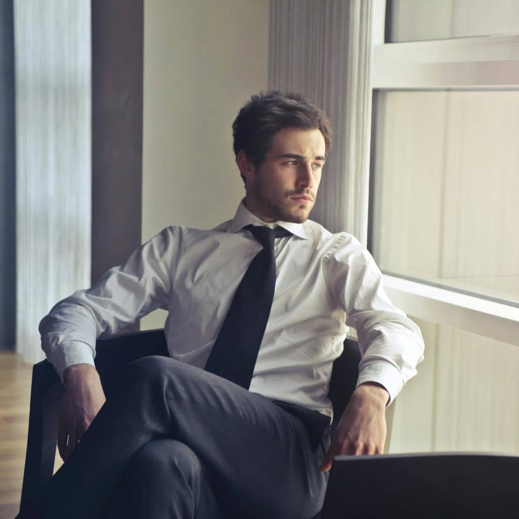 A man in a white shirt and black tie looking out a window while sitting on a chair.