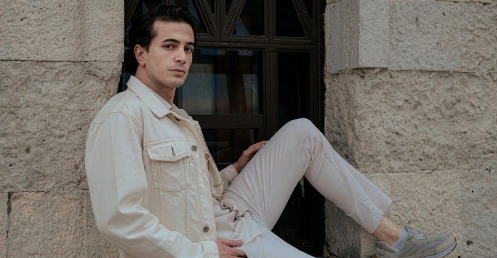 A man in a light-colored jacket and pants sitting on a ledge next to a building.