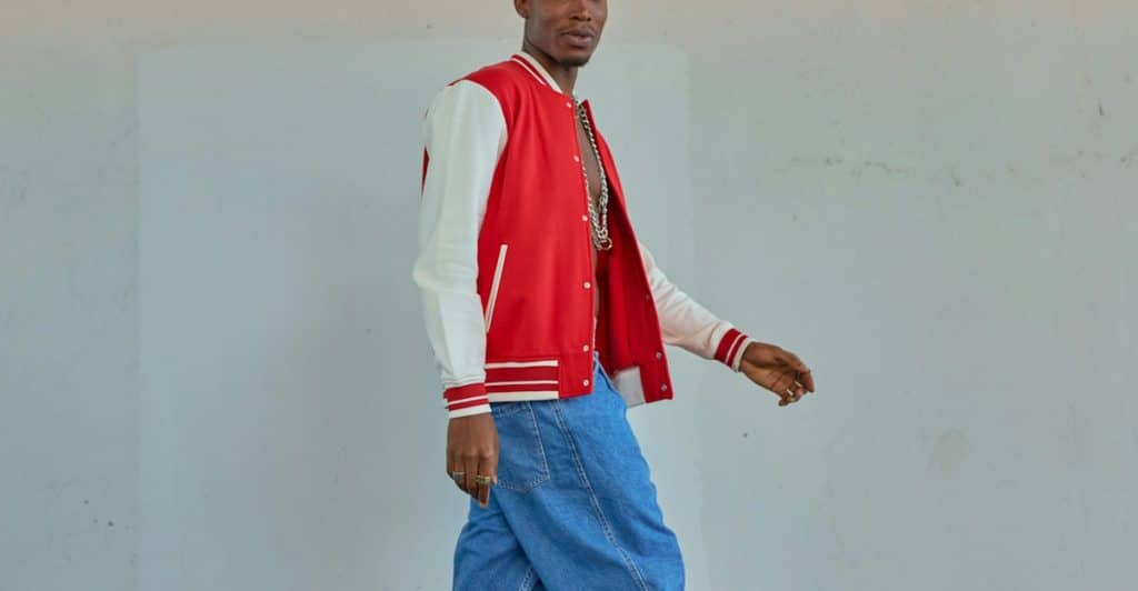 A man in a red and white varsity jacket and loose-fitting blue jeans is walking with his pants sagging low on his hips.