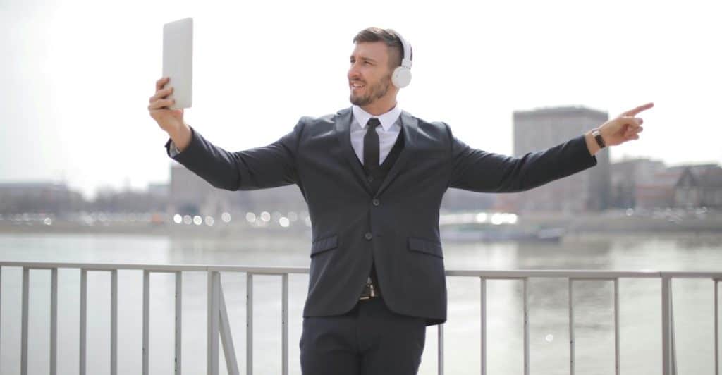 A man in a suit and headphones holding a tablet up, with his other arm extended pointing, standing outdoors near water and buildings.
