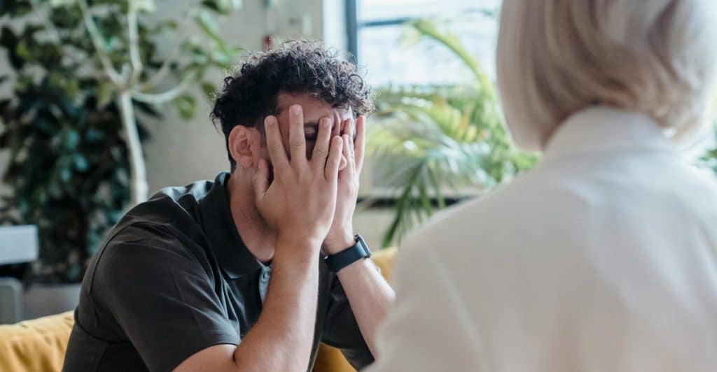 A man seated on a couch with his hands covering his face, while a therapist sits opposite him.