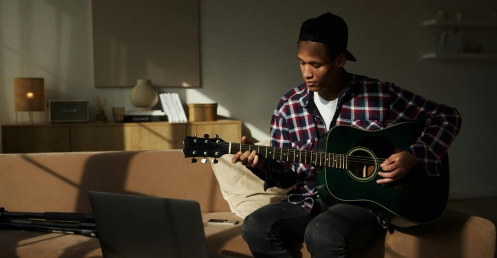 A man sitting on a couch with an acoustic guitar, looking at a laptop, possibly watching an online lesson.