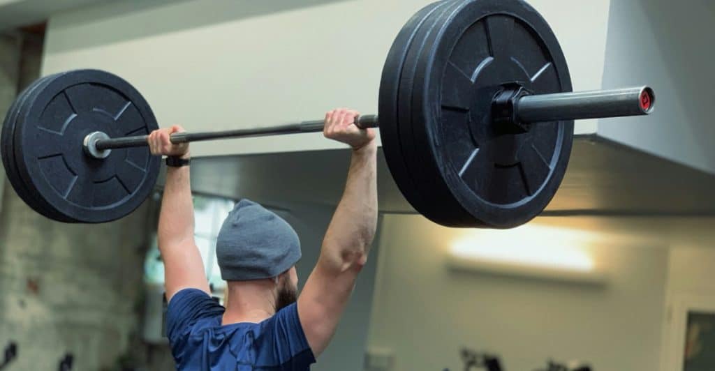 A man from behind, wearing a blue t-shirt and a grey beanie, is lifting a heavy barbell with large black weights overhead in a gym setting.