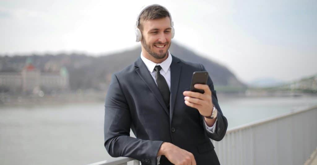 A man in a suit wearing headphones and smiling while looking at his phone outdoors.