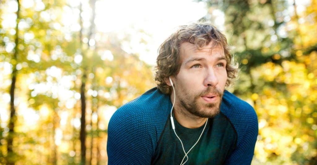 A man with curly hair and earbuds in his ears, looking up and slightly to the side in a forest setting with sunlight filtering through the trees.