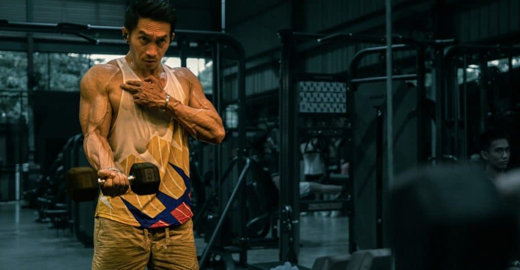 A muscular man in a white tank top and patterned shorts is holding a dumbbell and looking at his reflection in a gym mirror while performing repetitions.