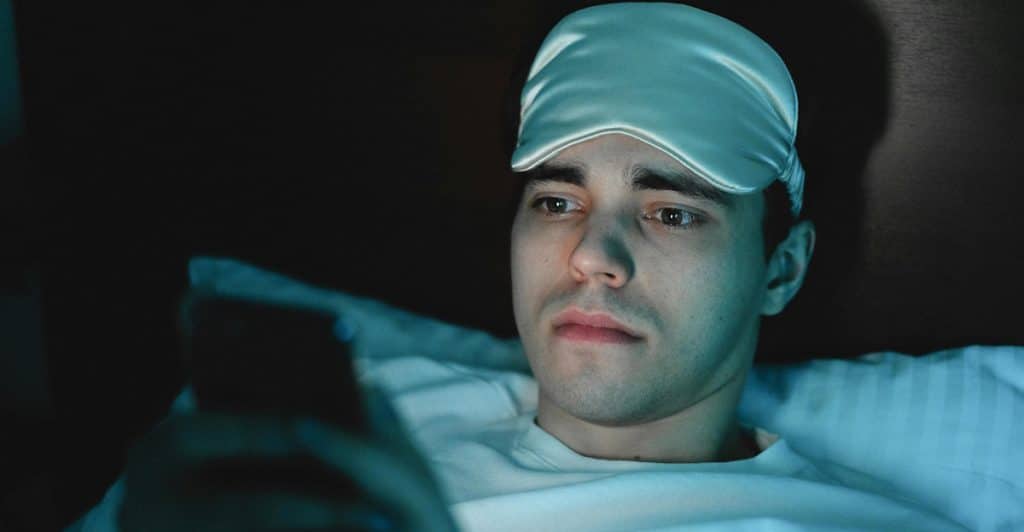 A man wearing a white sleep mask is looking at a phone in bed.