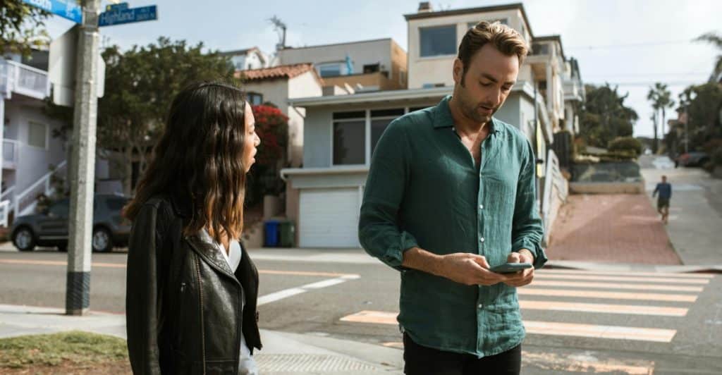 A man looking at his phone on a city street while a woman stands next to him, looking concerned.