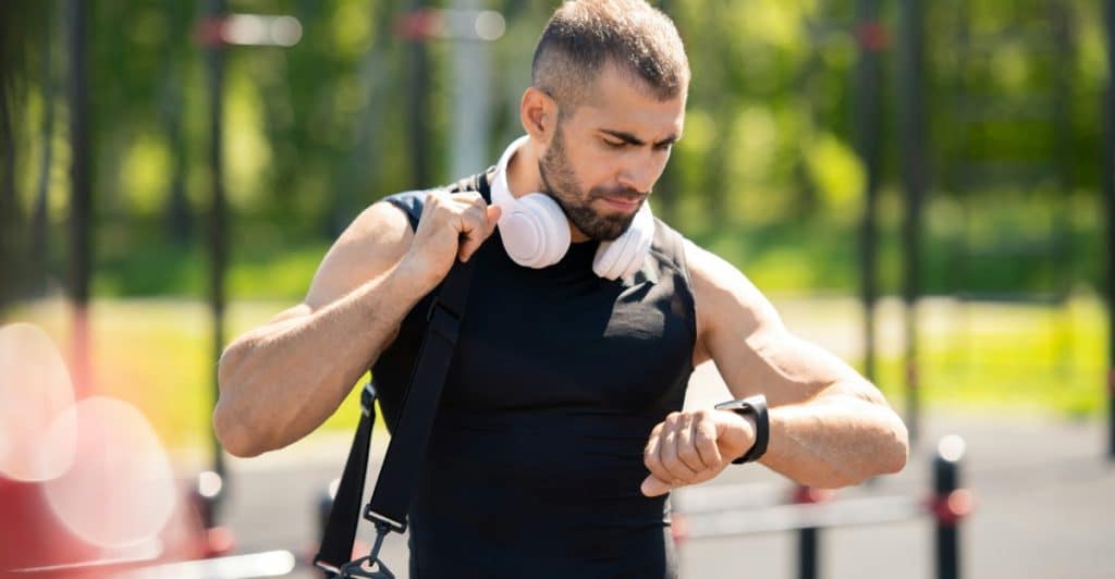 A fit man with headphones around his neck is outdoors, looking down intently at the smartwatch on his left wrist.