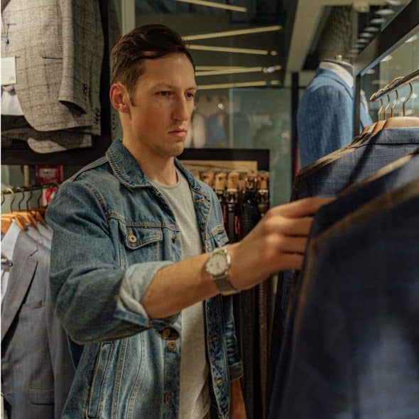 A man in a denim jacket is looking at suits hanging on a rack in a store.