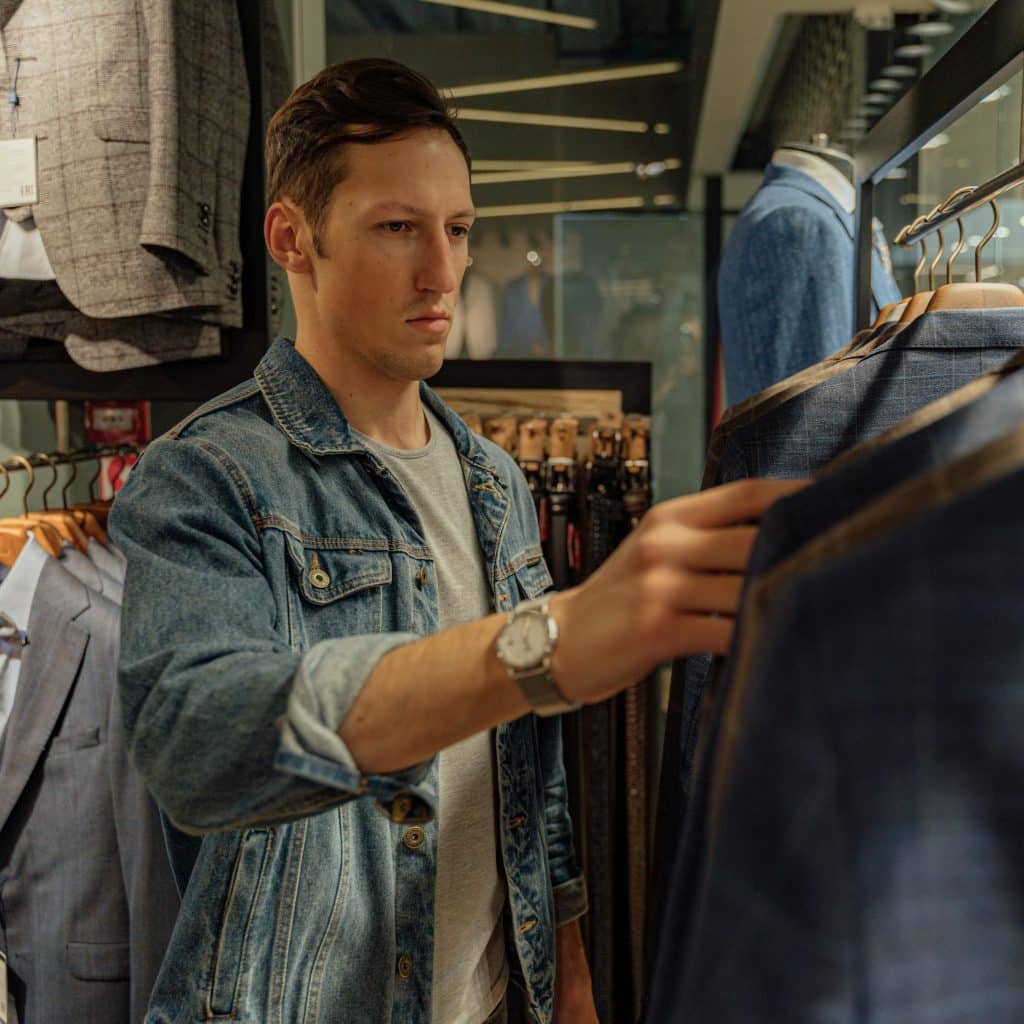 A man in a denim jacket is looking at suits hanging on a rack in a store.