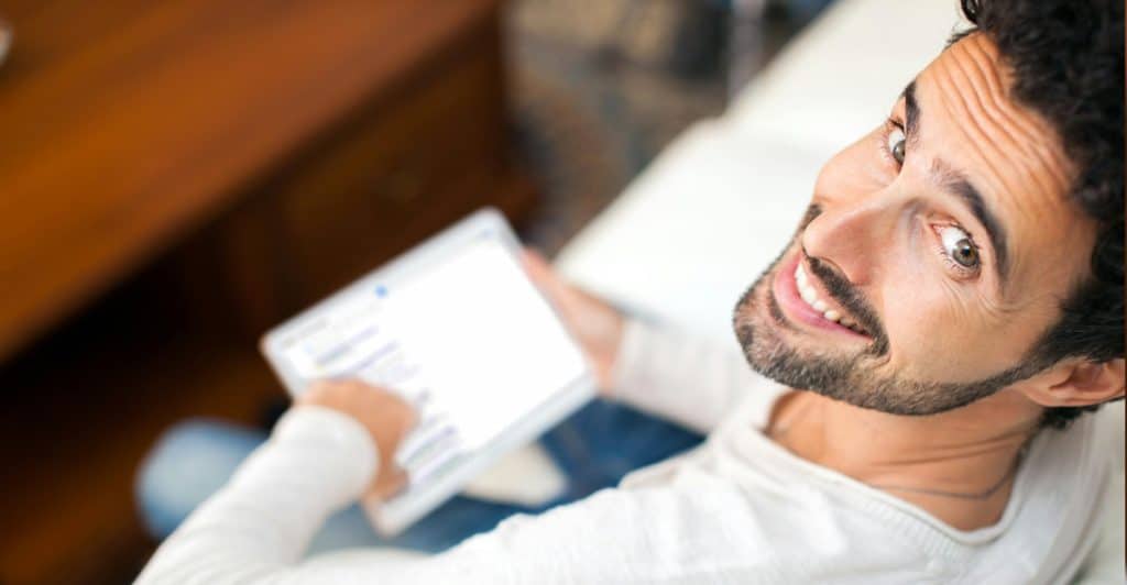 A man with dark curly hair and a beard is lying on a white couch, looking up and smiling at the camera while holding a white tablet.
