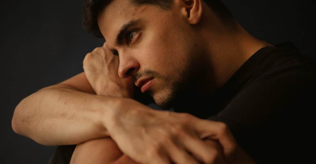 A man with short dark hair and a beard, looking down and away from the camera with his arms crossed over his knees.