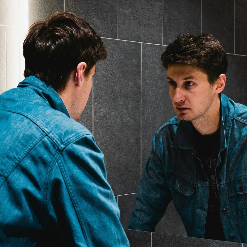 A man in a denim jacket looking intently at his reflection in a mirror.