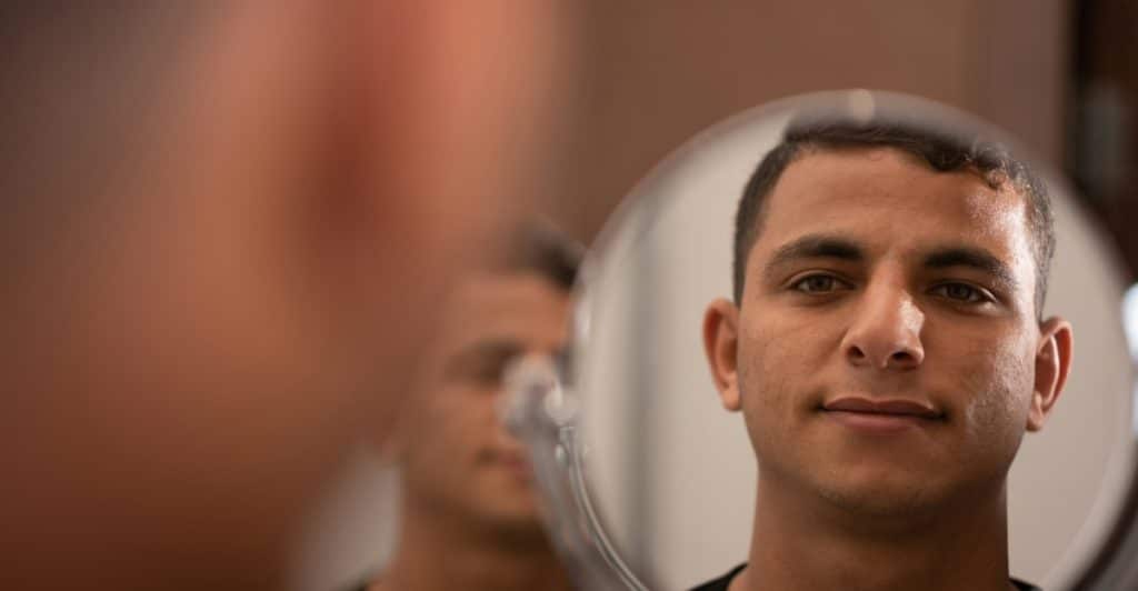 A man's face reflected in a small, round mirror, with a blurred reflection of his face in the background.