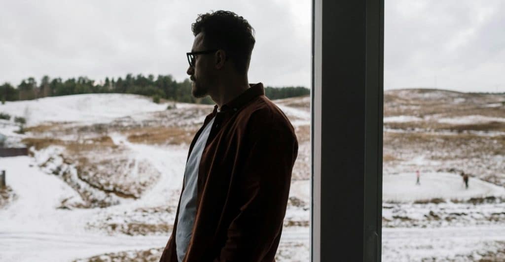 A man standing and looking out a window at a snowy landscape.