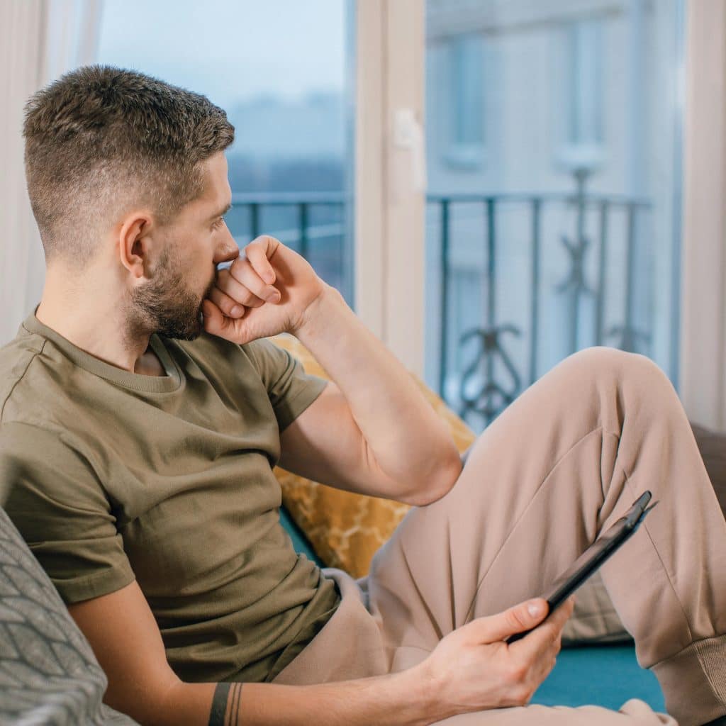 A man in a green shirt and beige pants sitting on a couch, looking out a window with a pensive expression, holding a tablet in his right hand.