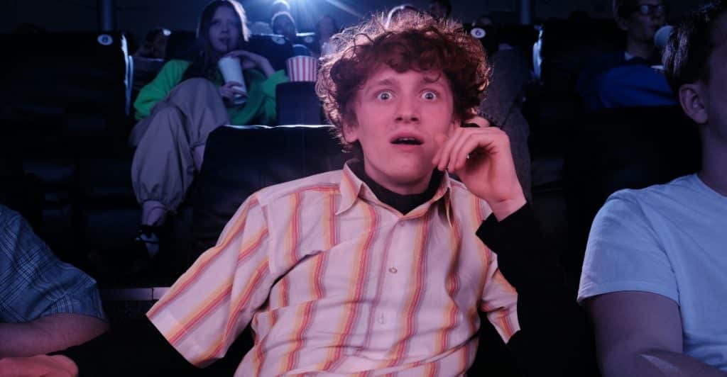 A man with curly hair looking shocked with his mouth open while sitting in a movie theater.