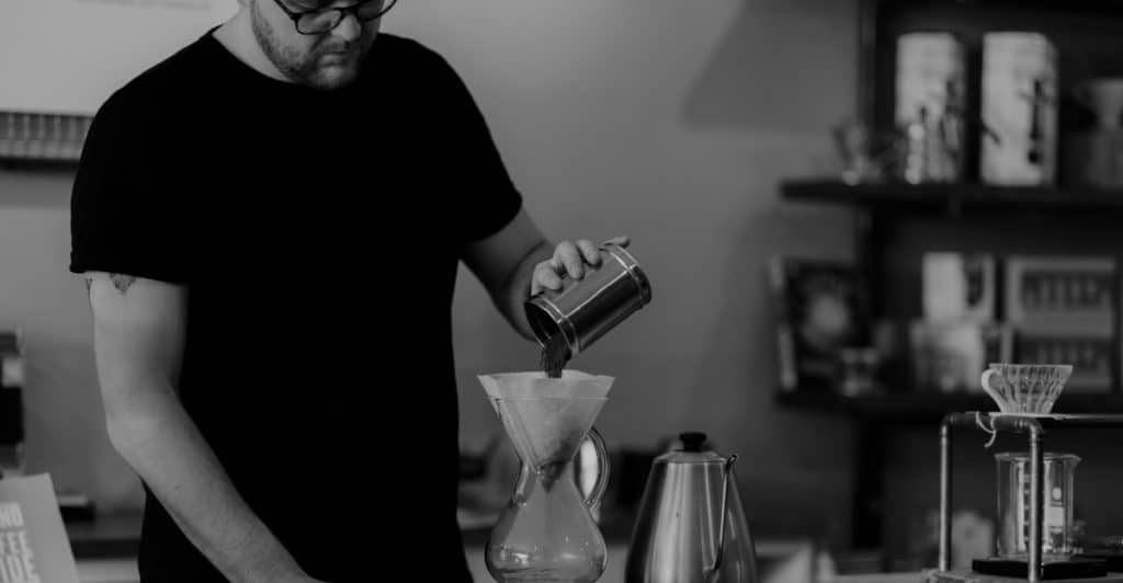 A man in glasses and a t-shirt pouring water from a metal pitcher into a pour-over coffee maker, set on a scale on a rustic counter, surrounded by coffee equipment.