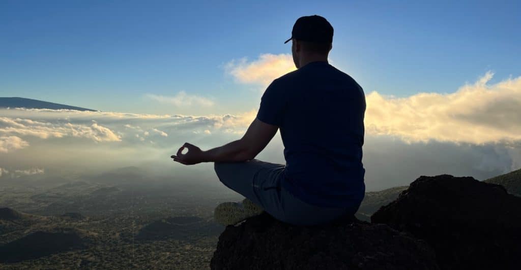 A man in a dark t-shirt and baseball cap is sitting in a lotus position on a rocky mountaintop, facing away from the viewer towards a cloudy sky with a bright horizon.