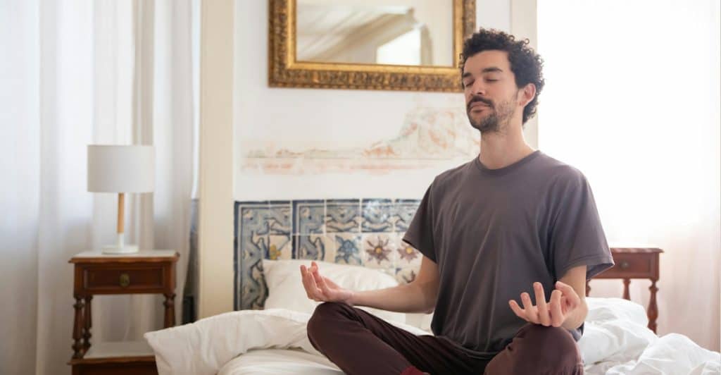 A man with dark, curly hair sitting in a meditative pose on a bed, with his eyes closed and hands resting on his knees.