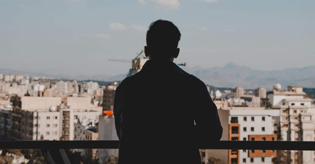 A silhouette of a man standing on a balcony overlooking a cityscape with mountains in the background.