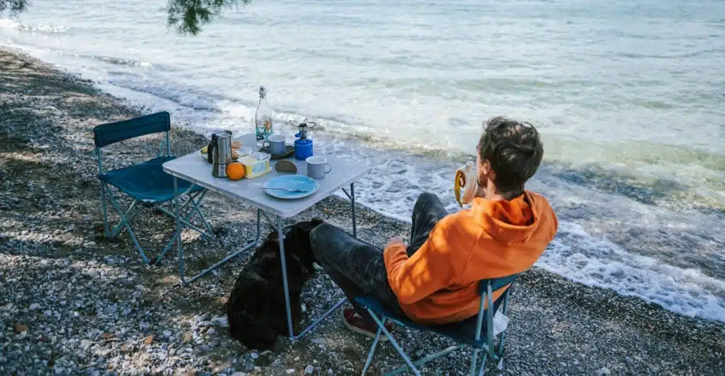 A man in an orange hoodie eating a banana while relaxing by a table set for breakfast on a pebbled beach near the sea.