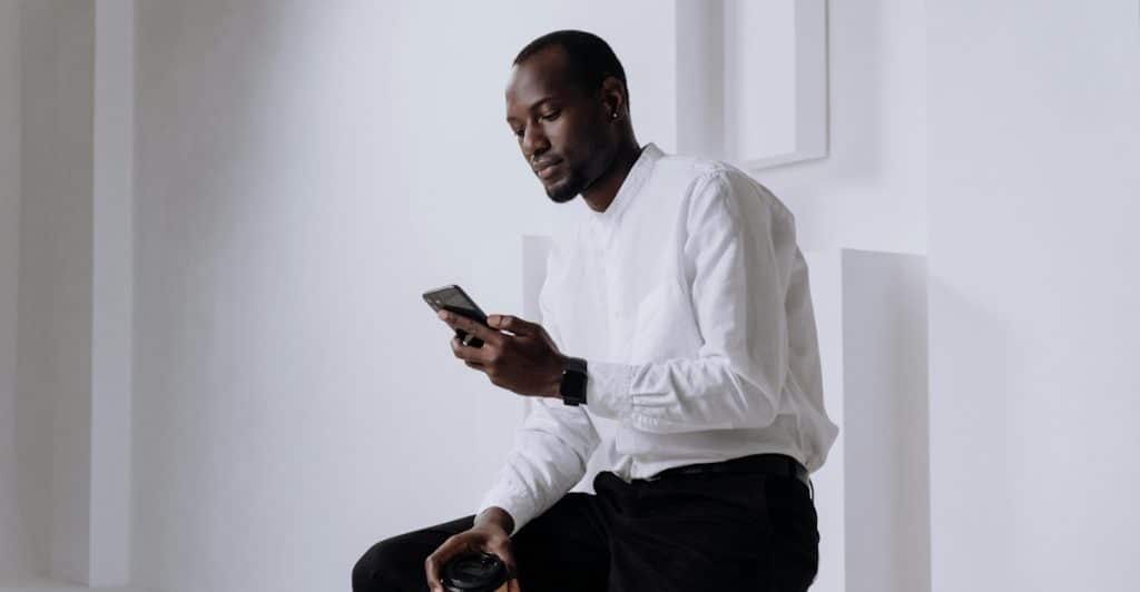 A man sitting on a stool, looking at his phone and holding a coffee cup.