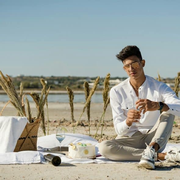 A man with sunglasses, wearing a white shirt and khaki pants, sitting on a white blanket on a beach, holding a glass and a bottle of wine, with a cake nearby.