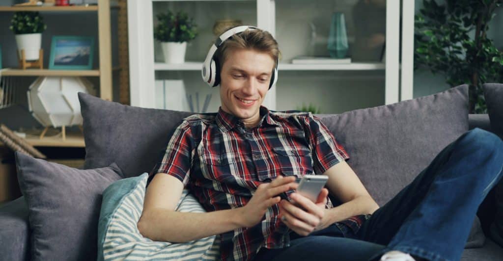 A man sitting on a couch wearing white headphones and holding a smartphone, looking at the screen and smiling.