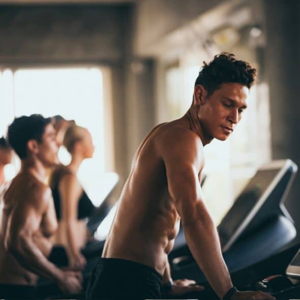 A shirtless man with dark, curly hair is looking down at the console of a treadmill he is standing on, with other people and treadmills visible in the blurred background of a gym.