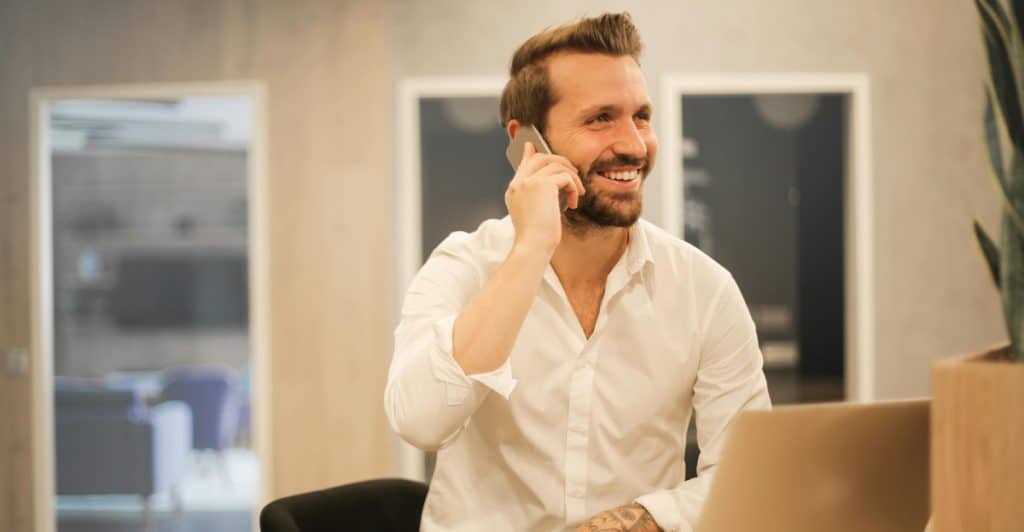 A man in a white shirt smiling while talking on a phone, sitting at a desk with a laptop and a cup of coffee.