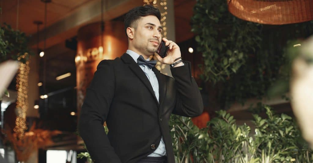 A man in a dark suit with a bow tie is standing in what appears to be a restaurant or cafe, smiling while holding a black smartphone to his ear with his left hand.