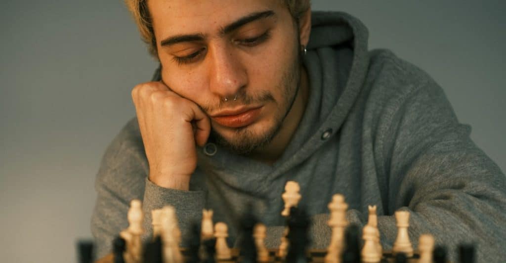 A man with blond hair and a nose ring, resting his head on his hand while looking at a chessboard.