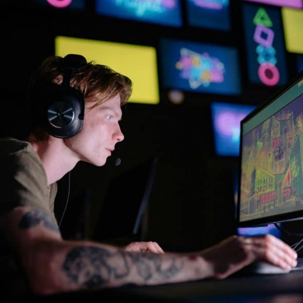 A man with headphones focused on a computer screen in a dimly lit room, with multiple colorful screens in the background.