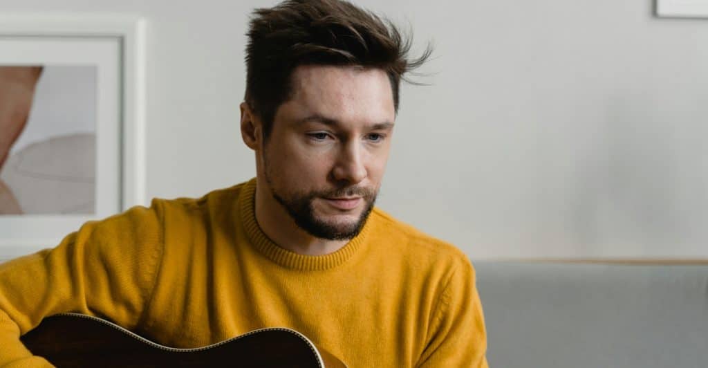 A man with a beard wearing a yellow sweater, playing an acoustic guitar.