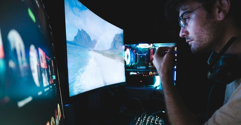 A man wearing glasses and headphones is seated in front of a large curved gaming monitor, with a glowing desktop PC visible to his right.