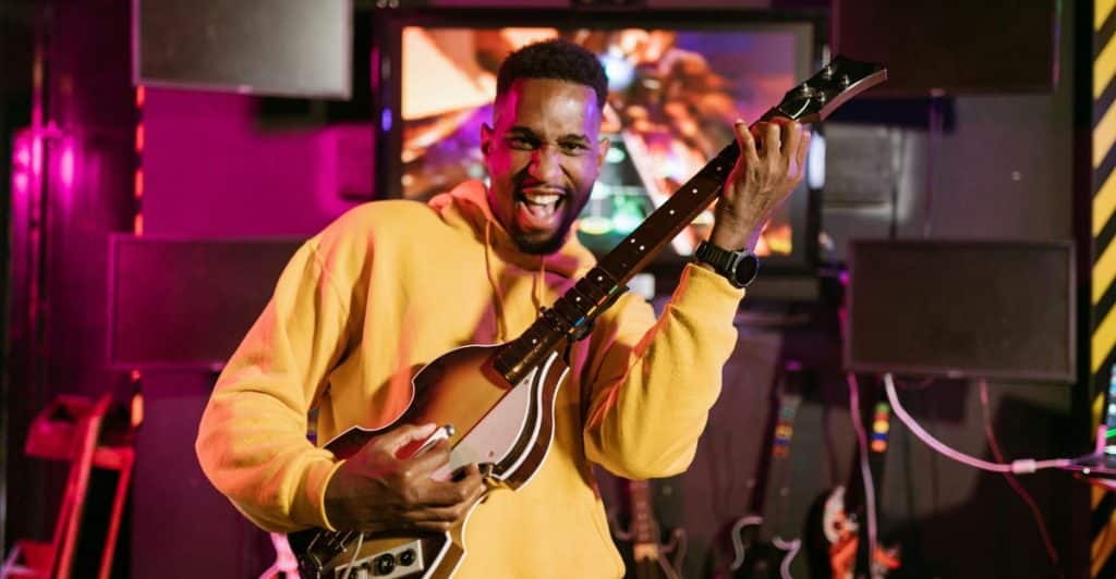 A man with a beard smiling and holding a virtual guitar, wearing a yellow hoodie, with screens and equipment in a room with pink and purple lighting.