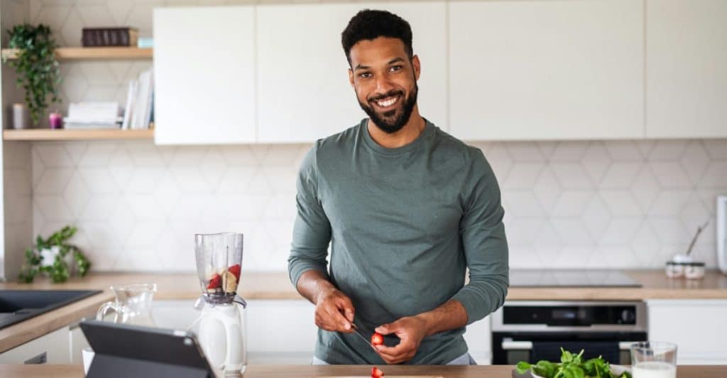A man smiling and looking at the camera while standing in a modern kitchen, preparing food with a blender and bowls of ingredients on the counter.