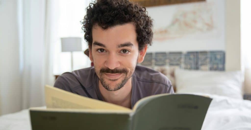 A man with curly hair and a mustache looking up from a book he is reading in bed.