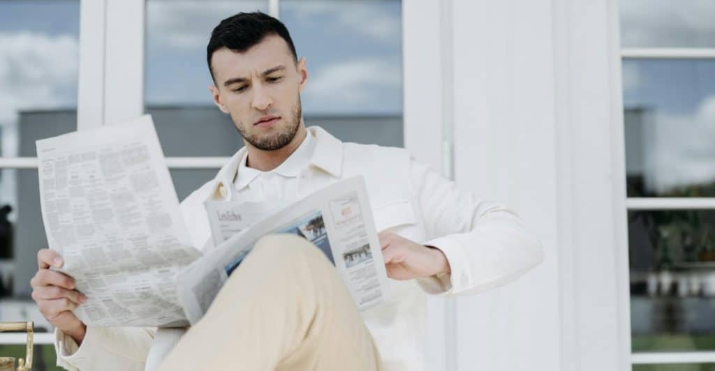 A man in a white jacket and light-colored pants sitting and reading a newspaper outdoors.