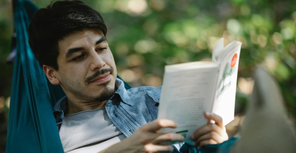 A man lying in a green hammock outdoors, engrossed in reading a book.