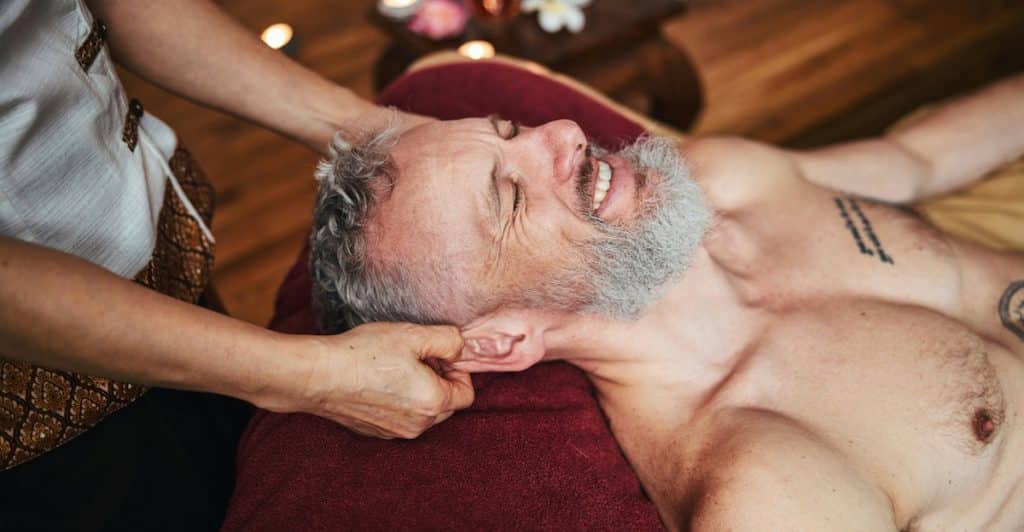 A man with a beard is smiling while receiving a neck massage from a person whose hands are visible.