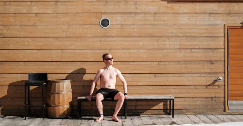 A man wearing sunglasses and shorts sits on a bench outdoors next to a wooden building.