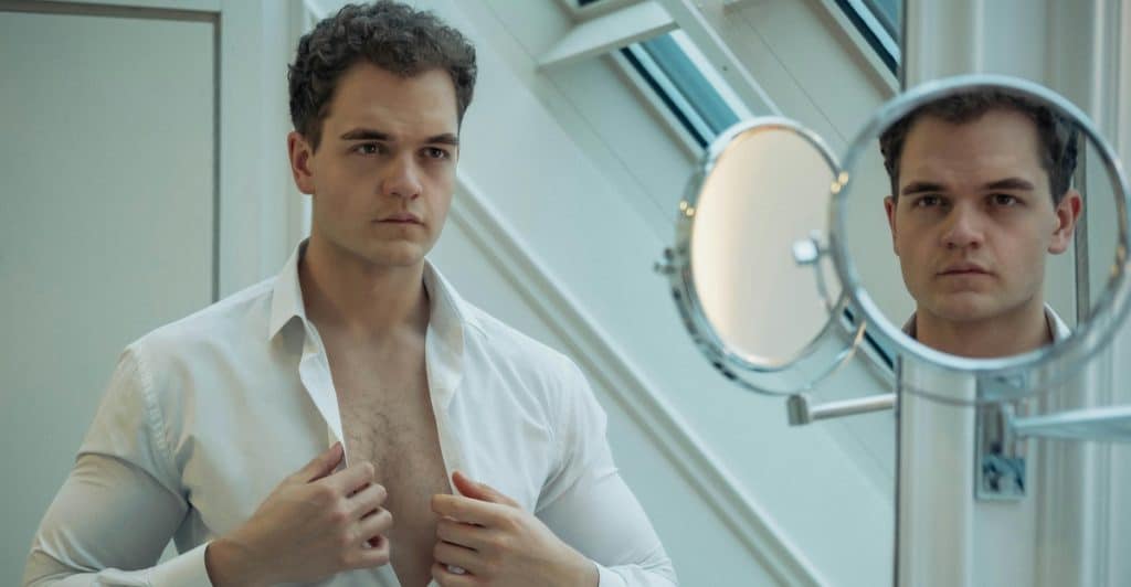 A man in a white shirt looking in a mirror and adjusting his shirt to reveal chest hair.